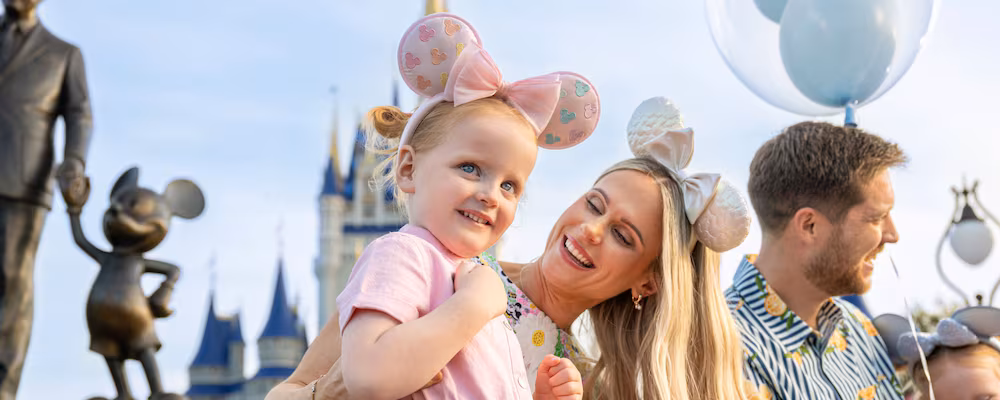 0507ZK_0478KCM_xak_adj-5x2 - SiDisney Smiling mother holding a little girl in pink Minnie ears with a castle and balloons in the background at a theme park.